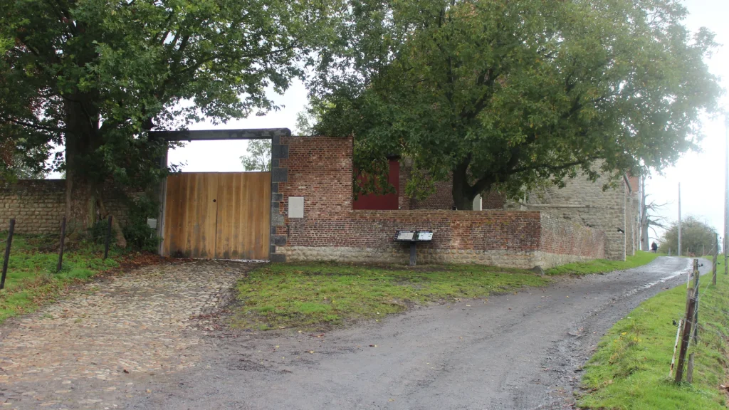 A rural scene showing an old brick building with a large wooden gate, partially open. a cobblestone path leads to the gate, flanked by lush green trees under overcast skies.