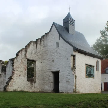 A small, historic white chapel with a gray roof alongside a partial stone wall under a cloudy sky, surrounded by green grass and a building to the right.