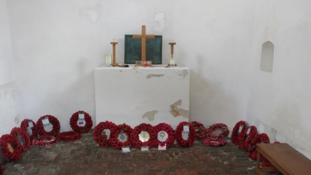 A simple church altar with a wooden cross and candles, surrounded by red poppy wreaths on the floor in a room with white walls and a brick floor.
