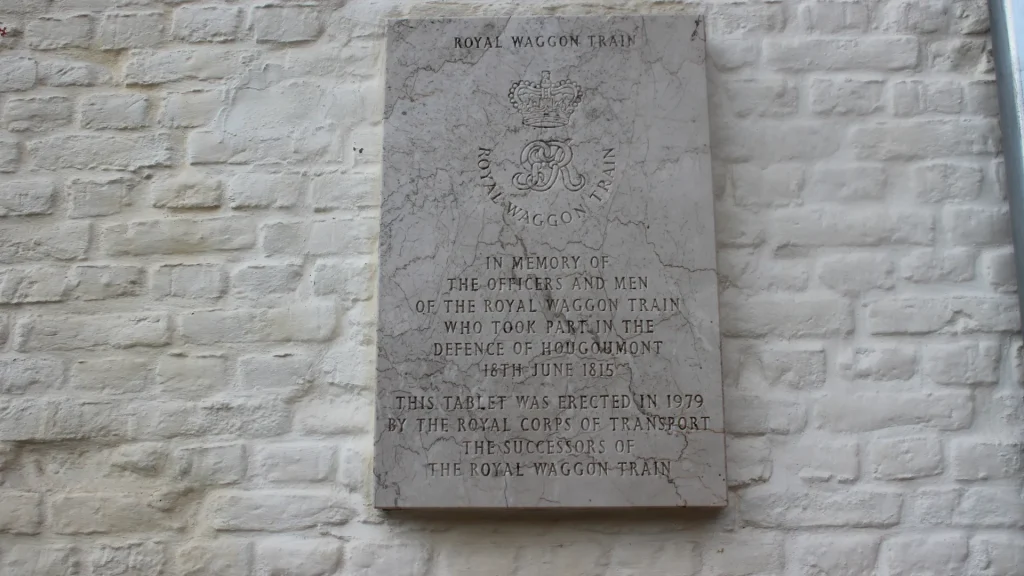 A commemorative plaque mounted on a white brick wall, dedicated to the officers and men of the royal waggon train for their defense at hougoumont on 16th june 1815, erected in 1919.