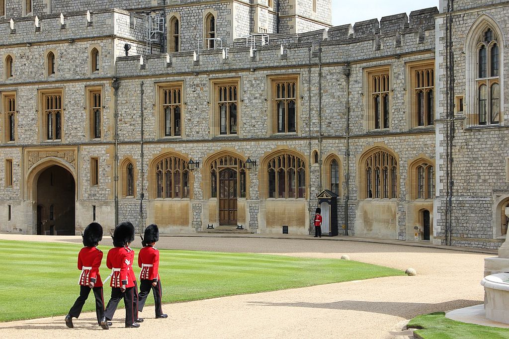 Guards in traditional red and black uniforms march in front of a historic castle with ornate stonework and large windows. one guard stands alone, watching.