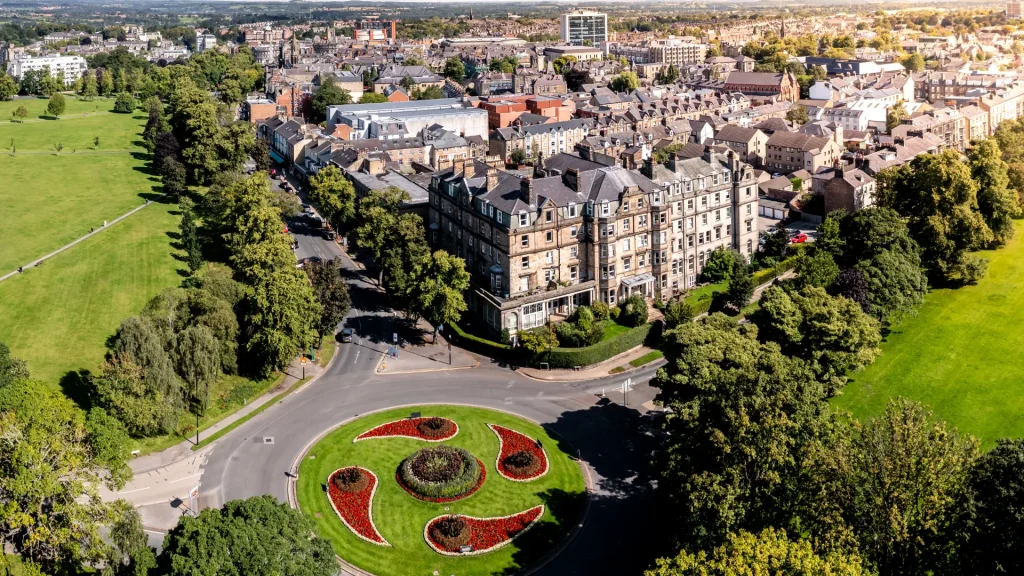 Aerial view of a vibrant, circular floral arrangement on a roundabout, surrounded by traditional multi-story buildings, with lush green parkland in the foreground and a quaint townscape in the background.