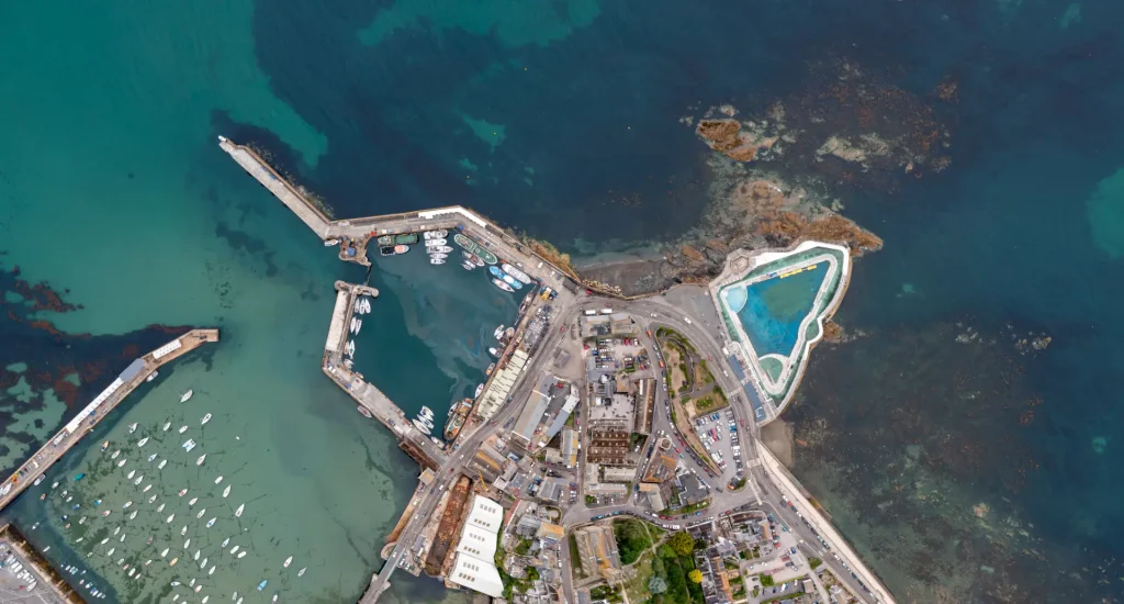 Aerial view of a coastal town with a marina, numerous boats, and a distinct turquoise swimming pool, surrounded by built-up areas and natural rocky coastline.