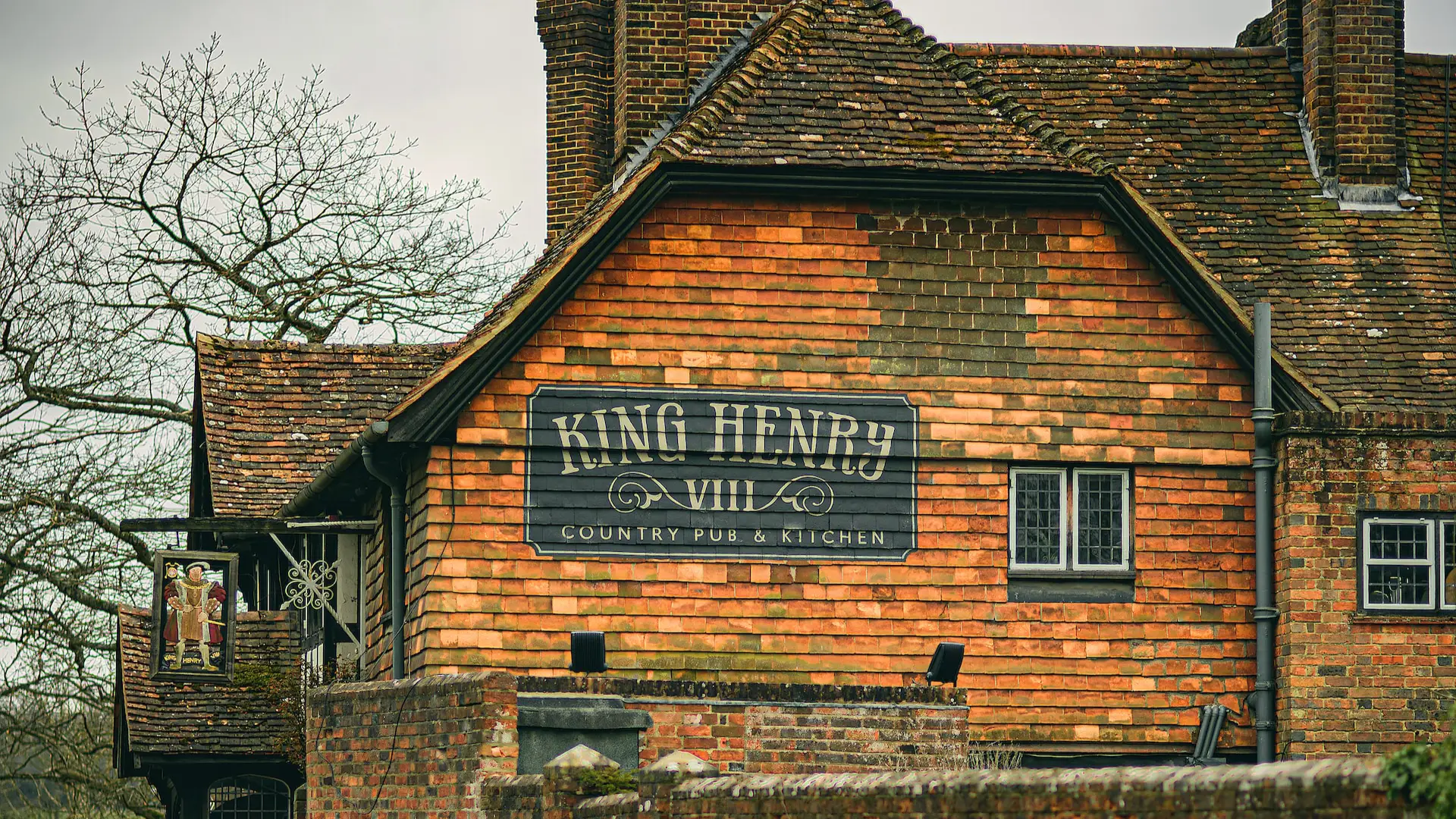 A quaint, brick-built country pub named "king henry viii" features a black sign with white lettering, set against a backdrop of lush trees. two people stand on a balcony adorned with flowers.