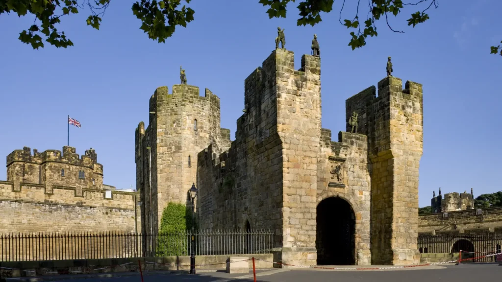A medieval stone castle with twin fortified gatehouse towers adorned with statues, under a clear blue sky. a british flag flutters atop one tower.