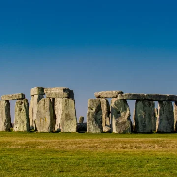 Stonehenge on a clear day, featuring the ancient stone circle with large standing stones set against a bright blue sky, with a few visitors visible around the monument.