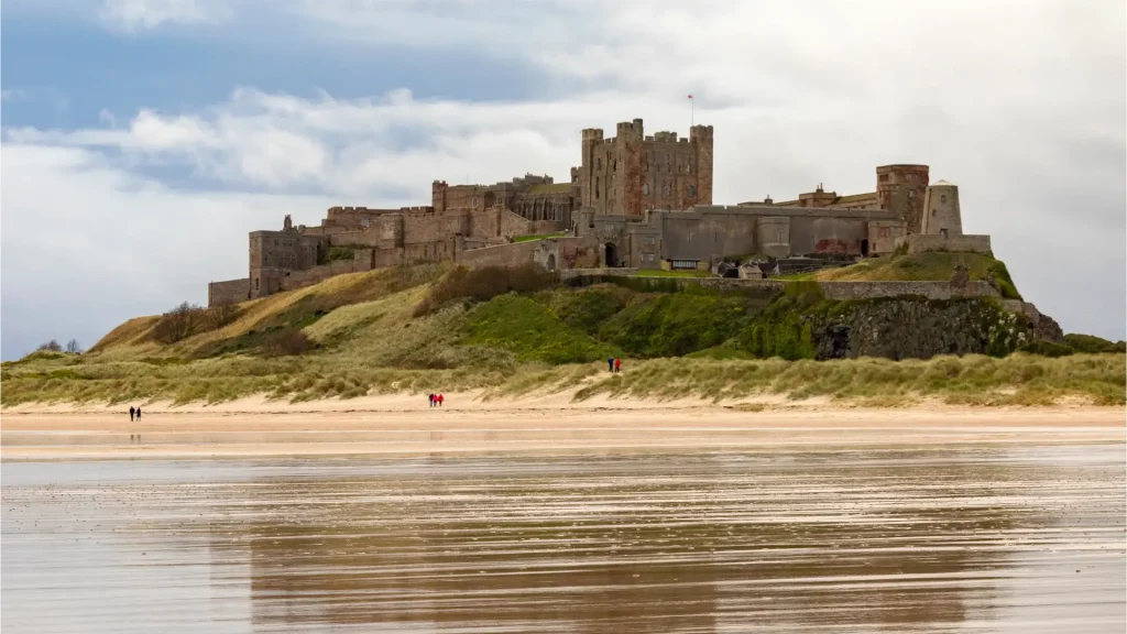 A majestic medieval castle situated on a grassy hill overlooking a sandy beach with a reflective wet surface, under a cloudy sky with people walking on the beach.