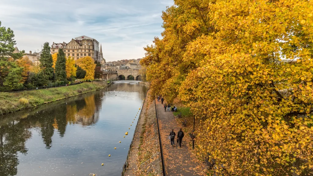 A scenic autumn view showing people walking along a tree-lined riverbank, with golden yellow leaves and a historic stone bridge in the background. the city's architecture looms over the serenity of the river.