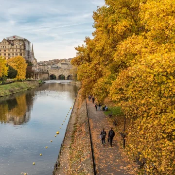 Autumn scene with people walking along a riverside path lined with golden yellow trees, a calm river on one side, and historic buildings in the background under a cloudy sky.