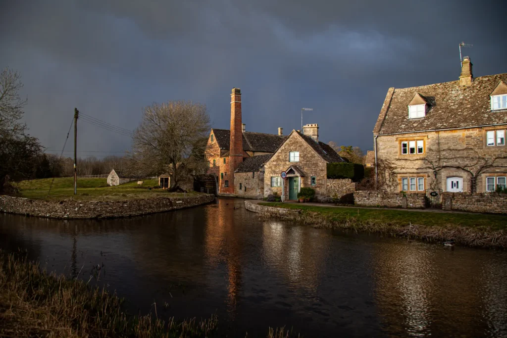 Old stone cottage with a tall chimney beside a calm river, under a stormy sky at twilight. well-manicured grass and a small footbridge are visible.
