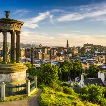 A panoramic view of edinburgh at sunset, showing the dugald stewart monument in the foreground and the cityscape, including historic buildings and green spaces, in the background.