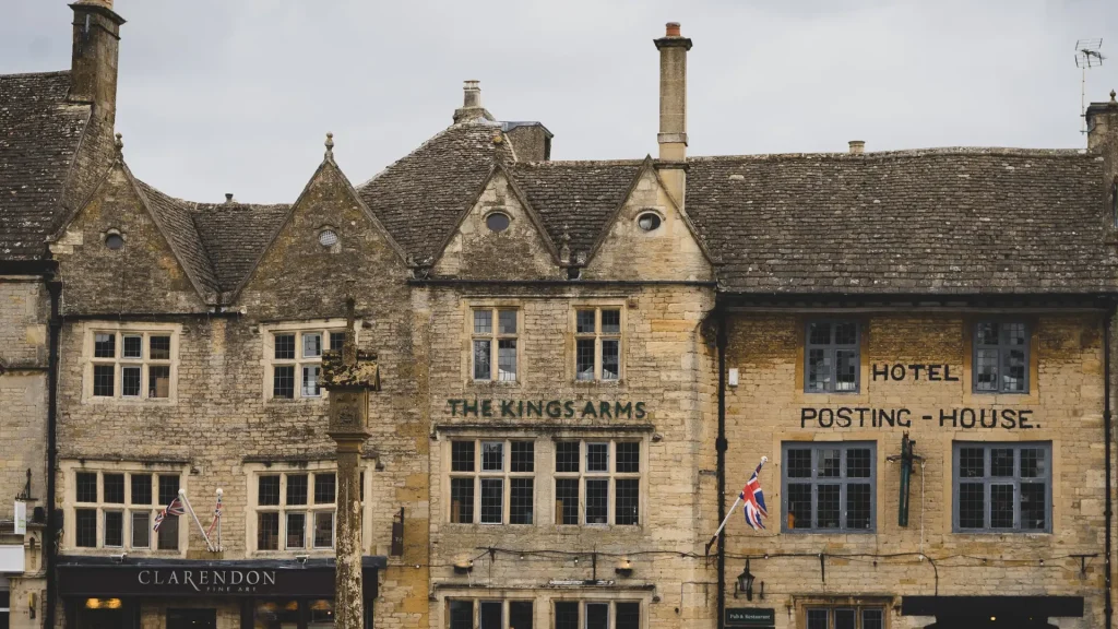Historic stone buildings with signs for "the kings arms" and "hotel posting house," featuring british flags, in a quaint town setting.