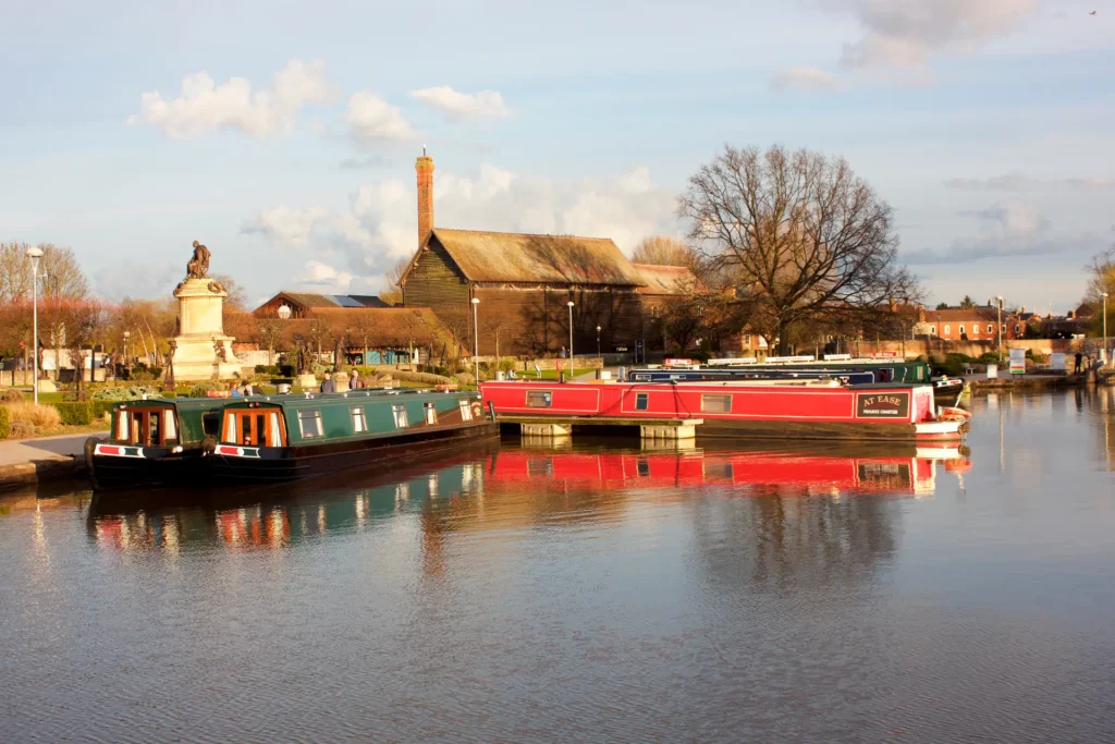Narrowboats moored on a calm river, with reflections visible in the water. a historic building and statue are seen in the background under a partly cloudy sky.