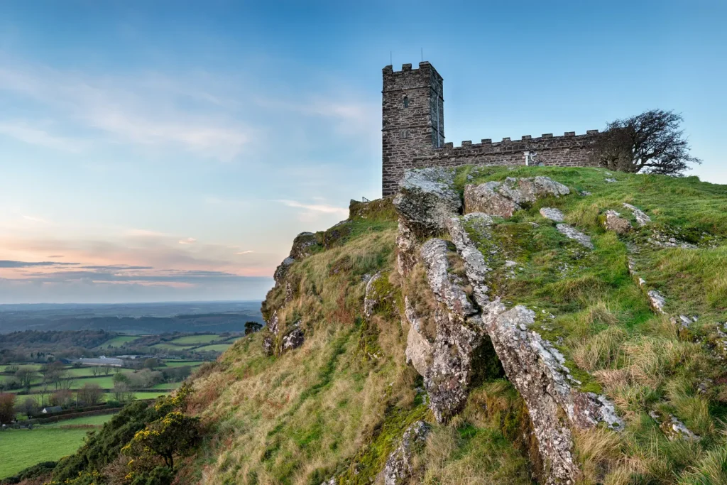 A scenic view of a stone tower atop a rugged hill during sunset, with sweeping views of the lush countryside below and a clear sky above.