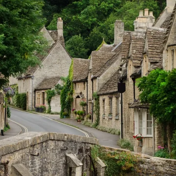 A picturesque village street lined with traditional stone houses, lush greenery, and a stone bridge in the foreground, embodying a quaint rural english charm.