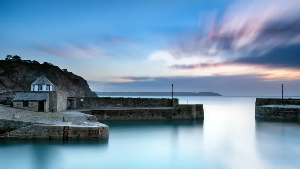 A serene harbor scene at dusk featuring a stone building at the dock, with smooth water reflecting the sky and long exposure streaky clouds moving over a silhouetted coastline in the distance.