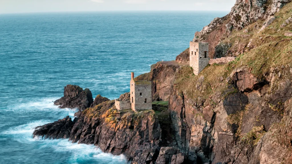 Old stone towers perched on a rugged cliff overlooking the ocean with waves crashing against the rocks below under a clear sky.