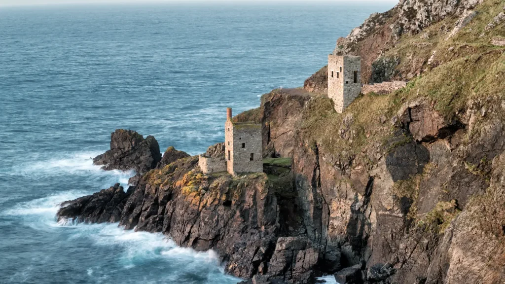 A rugged coastline featuring old stone structures on a cliffside overlooking tumultuous blue sea waves, under a clear sky.