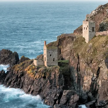 A rugged coastline featuring old stone structures on a cliffside overlooking tumultuous blue sea waves, under a clear sky.