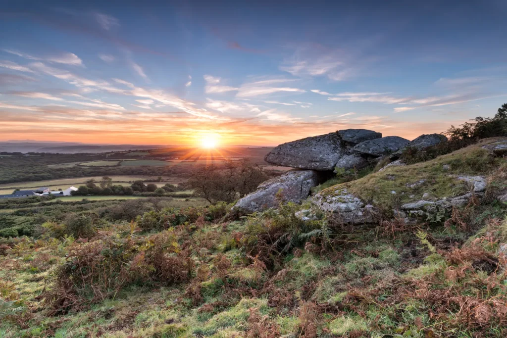 Sunrise over a picturesque landscape, featuring a rocky outcrop surrounded by lush greenery with a distant view of rolling hills and a serene sky.