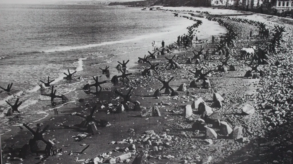 Historical black and white photo of a beach fortified with large anti-tank obstacles during wartime, crowded with troops and located adjacent to a tree-lined promenade.