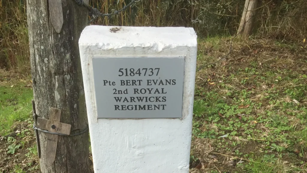 A white concrete boundary marker with a plaque displaying "5184737 pte bert evans 2nd warwicks regiment" next to a wooden post and greenery in the background.
