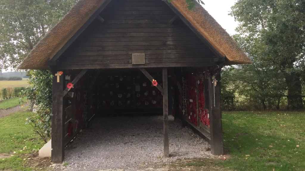 A traditional thatched roof shelter displaying an array of poppy wreaths and commemorative plaques on its wooden walls, located in a rural setting with visible trees and fields in the background.