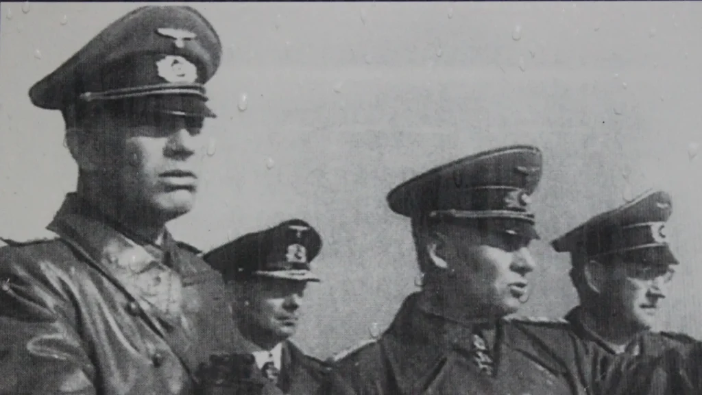 Black-and-white photo of four uniformed men with military caps, visible in profile, standing in line, with raindrops partially obscuring the image.