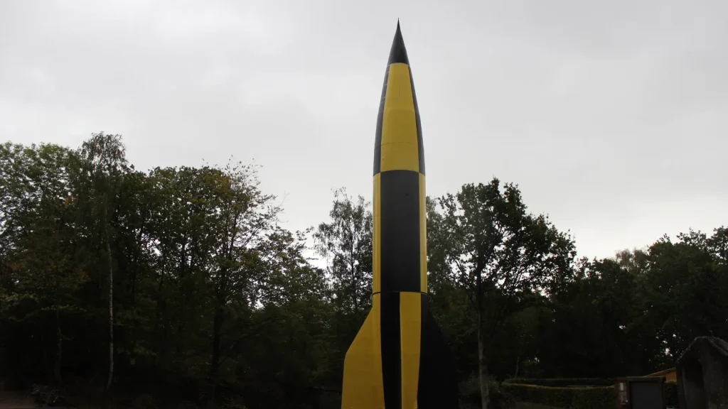 A large pencil-shaped sculpture stands tall against a cloudy sky, painted in alternating black and yellow stripes, surrounded by lush green trees.