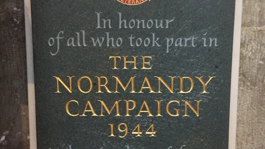 Close-up of a memorial plaque honoring participants of the normandy campaign in 1944, featuring golden text on a dark stone background.