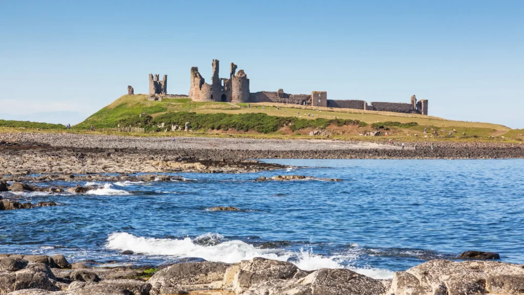 A picturesque scene of a historic castle with multiple towers, perched on a grassy hill overlooking a calm blue sea with rocky shoreline under a clear blue sky.