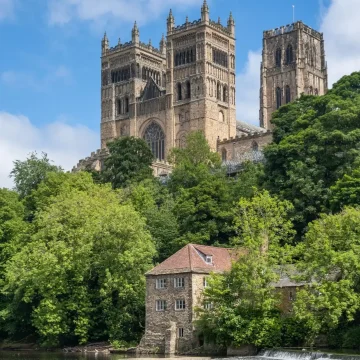 A majestic cathedral with twin towers overlooks a serene river, with a quaint stone house nestled among lush green trees in the foreground.