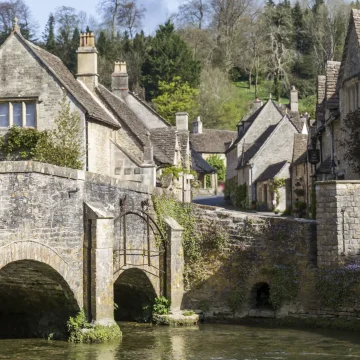 A quaint old stone bridge arching over a serene river in a picturesque village with traditional stone cottages surrounded by lush greenery under a clear sky.