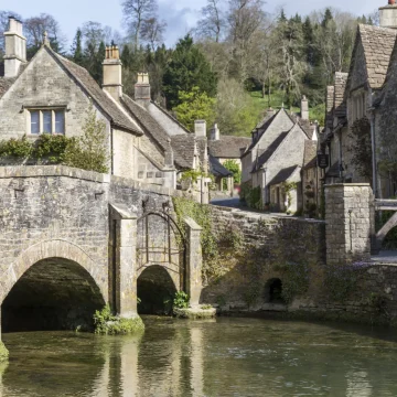 A quaint old stone bridge with two arches spans over a tranquil river, surrounded by traditional cottages in a picturesque rural setting.