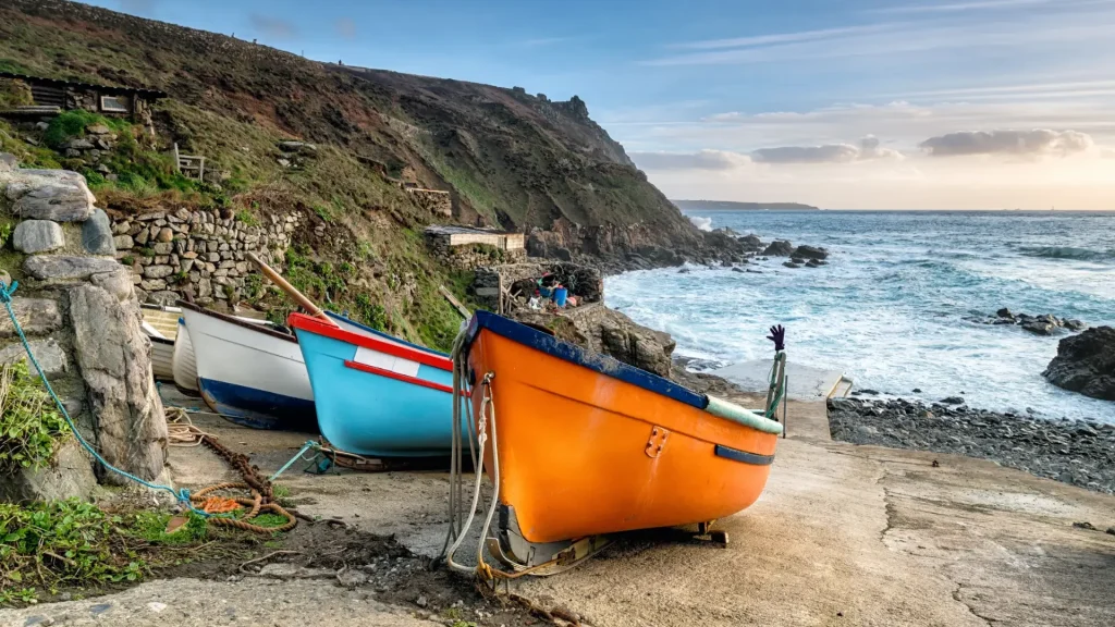 Three colorful boats on a rocky shore with turbulent sea waves and a rugged cliff under a cloudy sky.