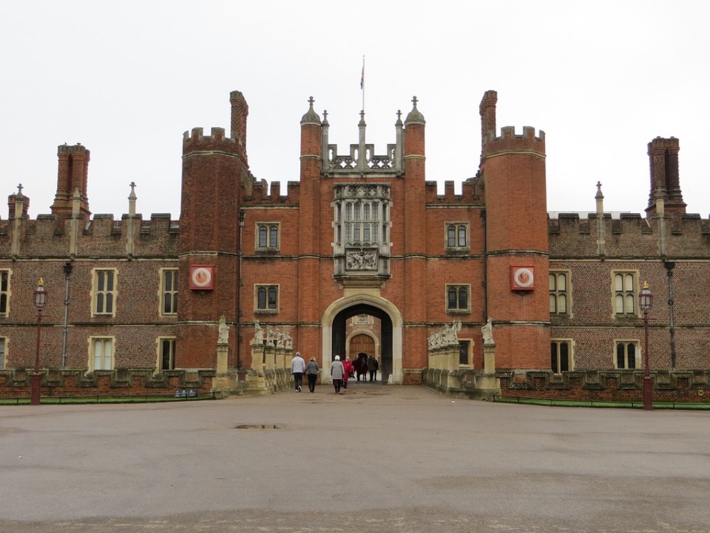 A group of people walking towards the grand entrance of a large historic red brick castle with ornate architectural details and turrets, under an overcast sky.