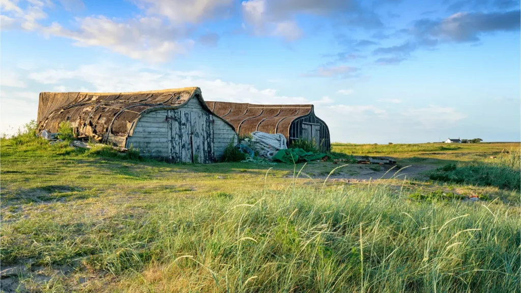 Two weathered boat sheds with curved roofs covered in black tar on a grassy field under a clear sky, with a covered boat on the side and a wide open landscape in the background.