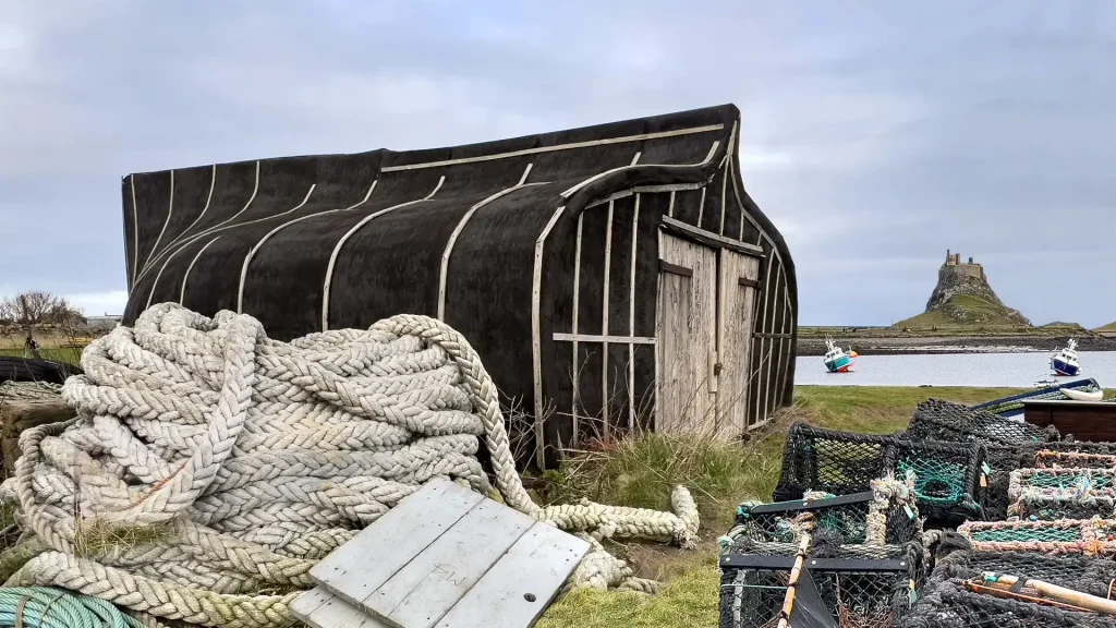 A rustic wooden boathouse with a curved roof, resembling an overturned boat, beside nautical equipment including ropes and nets, with a distant island visible under a cloudy sky.