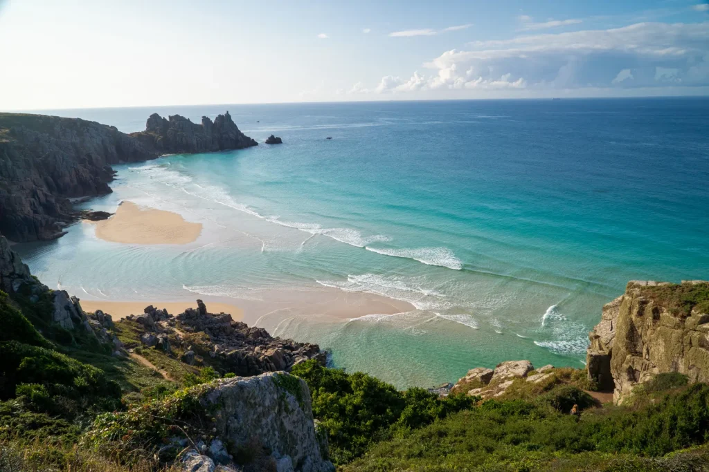 A serene coastal scene with turquoise waters gently washing onto a sandy beach flanked by rugged cliffs under a partly cloudy sky.