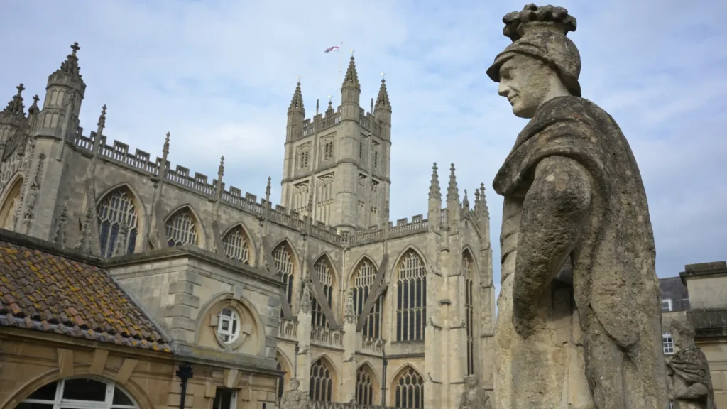 Stone statue of a robed figure overlooking the ornate gothic architecture of bath abbey, under a cloudy sky.