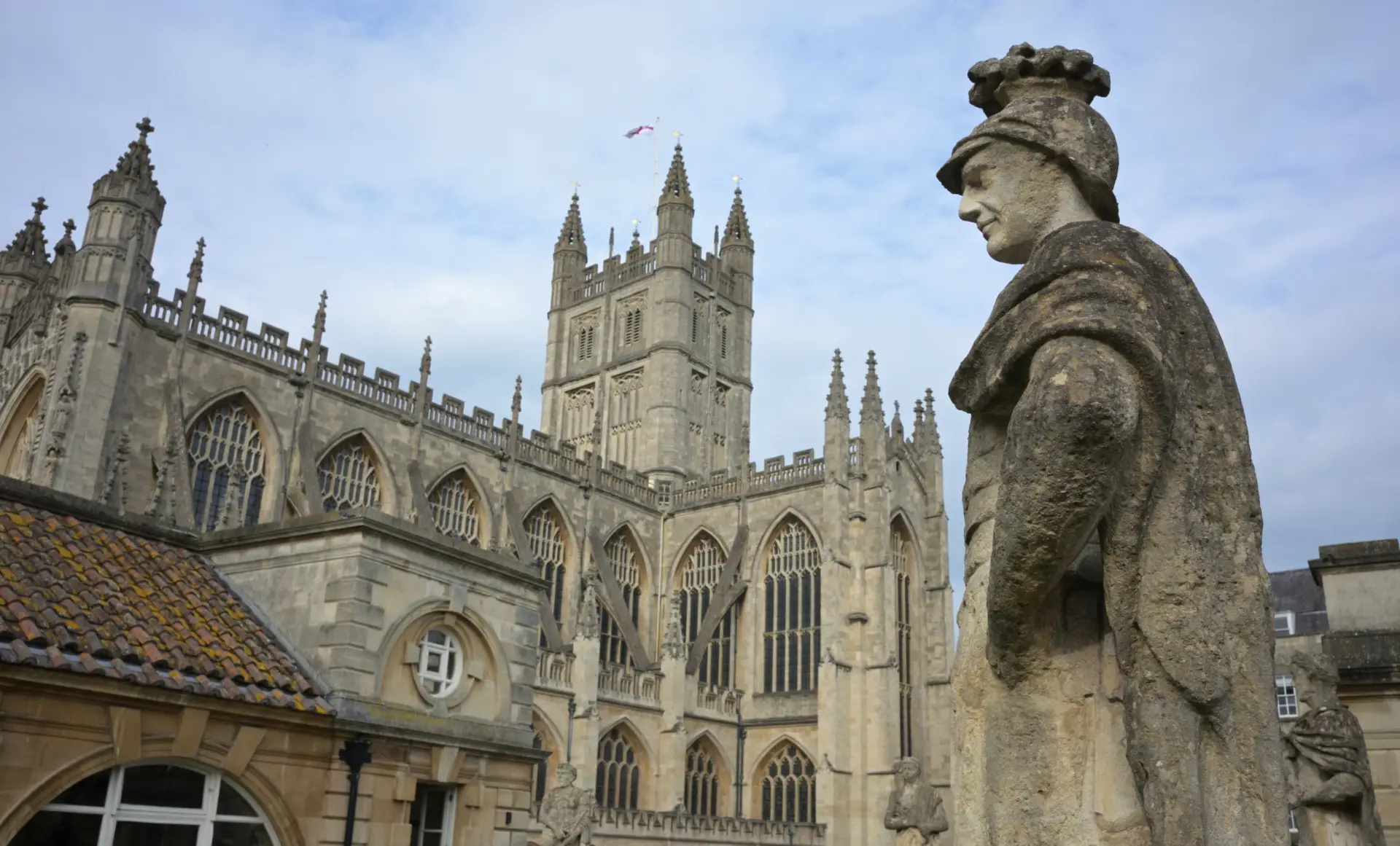 A stone statue of a historical figure in the foreground with the ornate architecture of a gothic cathedral, featuring spires and intricate facade details, in the background under a cloudy sky.