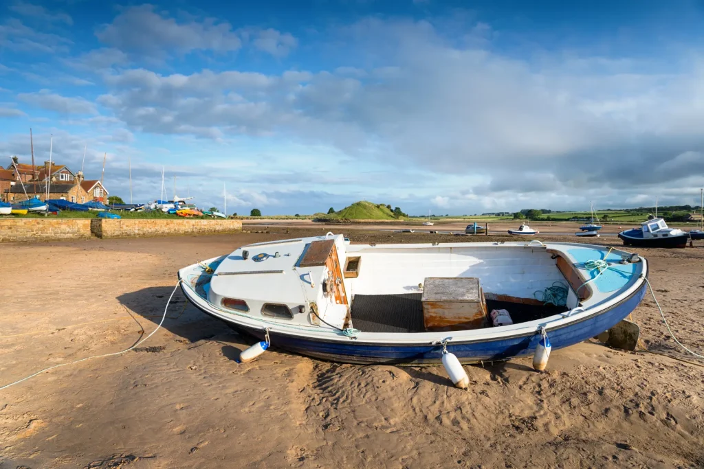 A weathered boat sits on a sandy shore with other boats and buildings in the background under a partly cloudy sky.