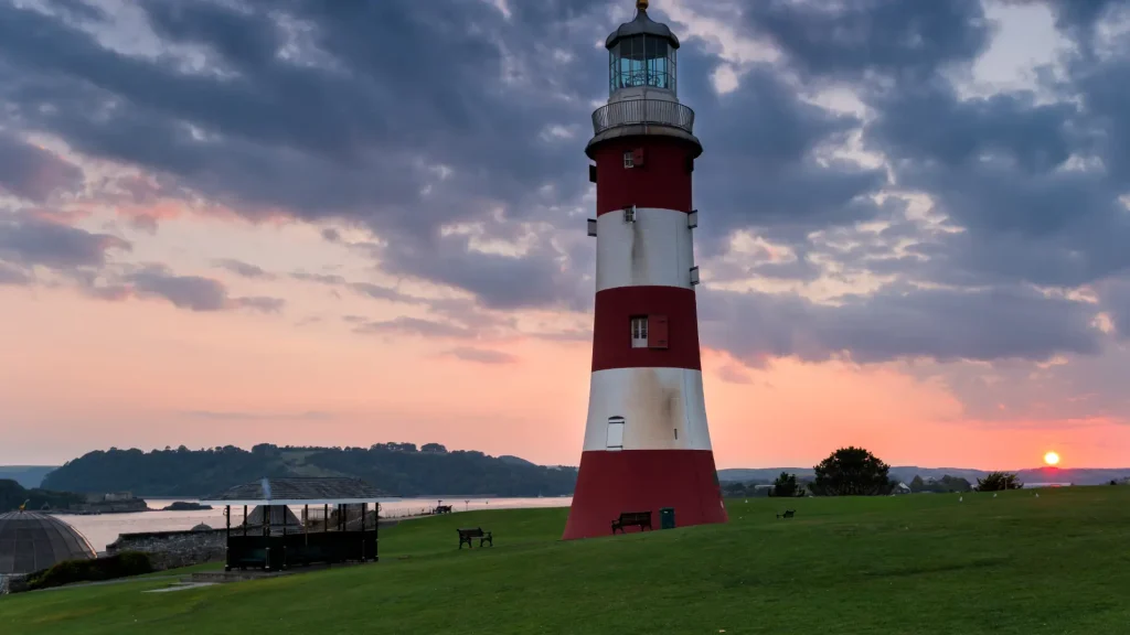 A striking red and white striped lighthouse stands on a grassy hill at sunset, with a colorful sky in the background and a serene body of water visible.