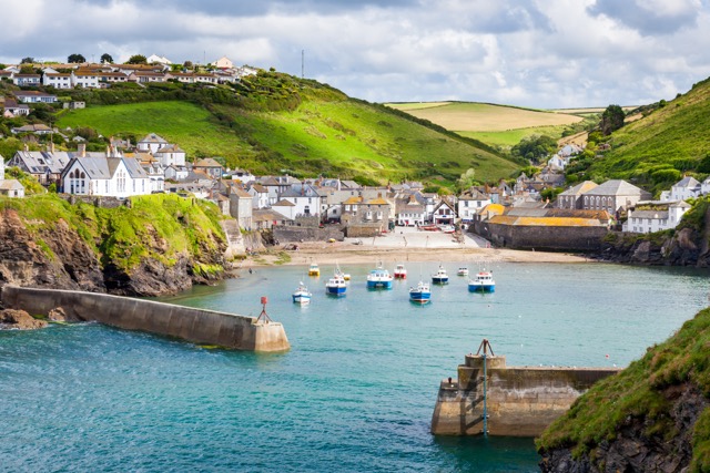 Picturesque coastal village with clustered houses along a hillside, a small harbor with boats, and a protective seawall under a partly cloudy sky.