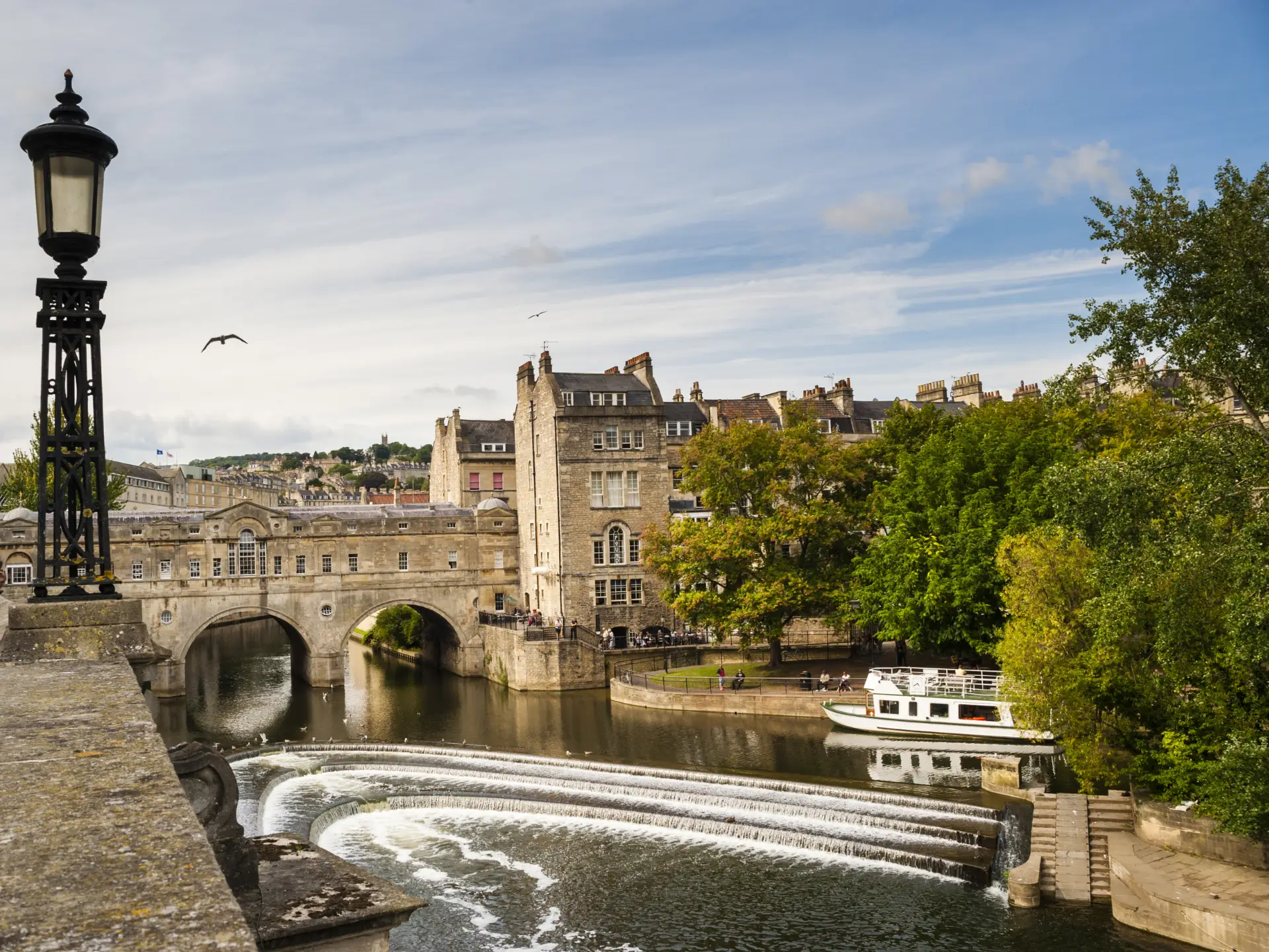 Historic pulteney bridge over the river avon in bath, england, with a beautiful cascading weir, a boat, and classic georgian architecture under a cloudy sky.