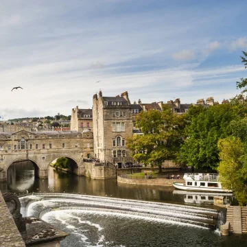 A scenic view of pulteney bridge and the horseshoe-shaped weir on the river avon in bath, with a boat cruising nearby and buildings lining the riverbanks under a cloudy sky.