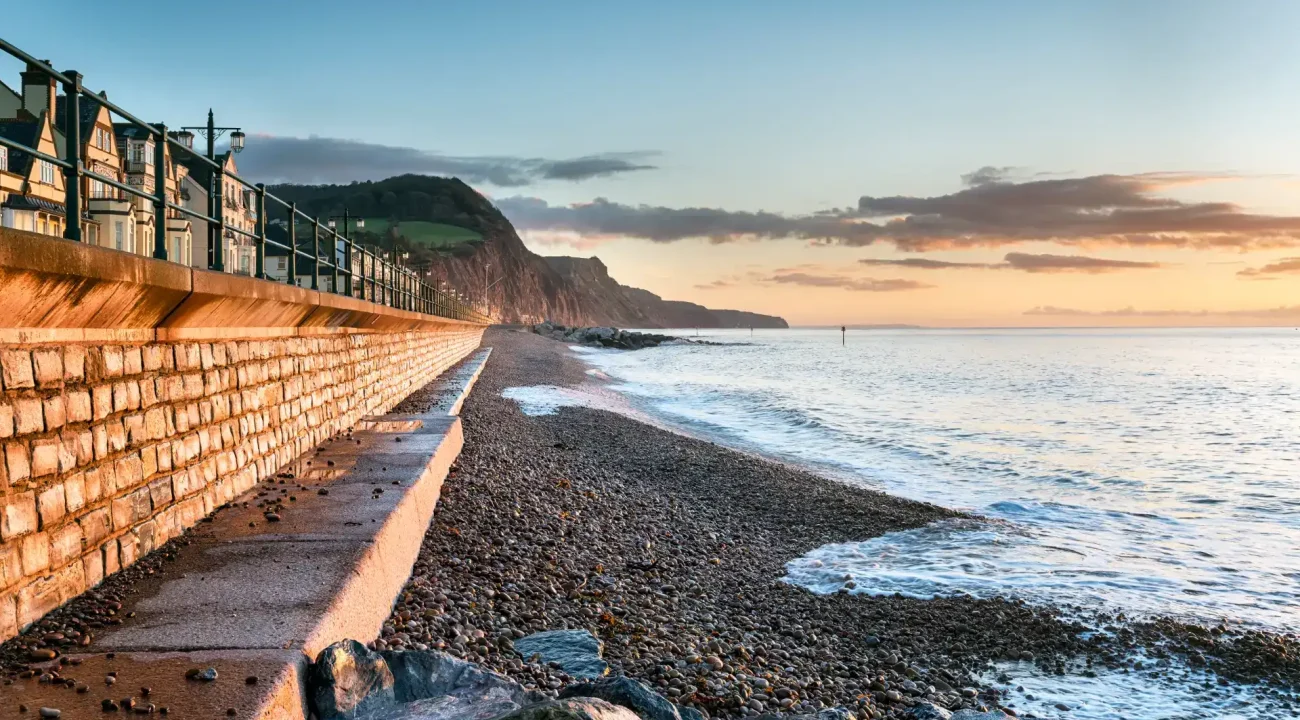 A serene coastline at sunrise featuring a pebble beach on the right, a long sea wall in the middle, and a hill extending into the distance on the left, under a clear sky.