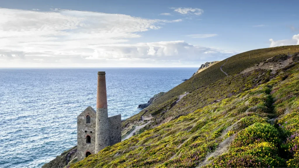 A scenic coastal landscape featuring a historic stone tower with a tall chimney, perched on a hillside covered with lush greenery and wildflowers, overlooking a bright blue sea under a partly cloudy sky.
