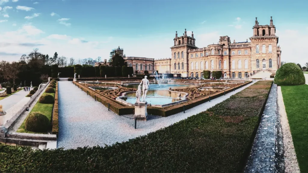 A panoramic view of a grand historic palace with elaborate architecture and multiple towers, surrounded by manicured gardens, sculpted hedges, and a central fountain featuring a statue.