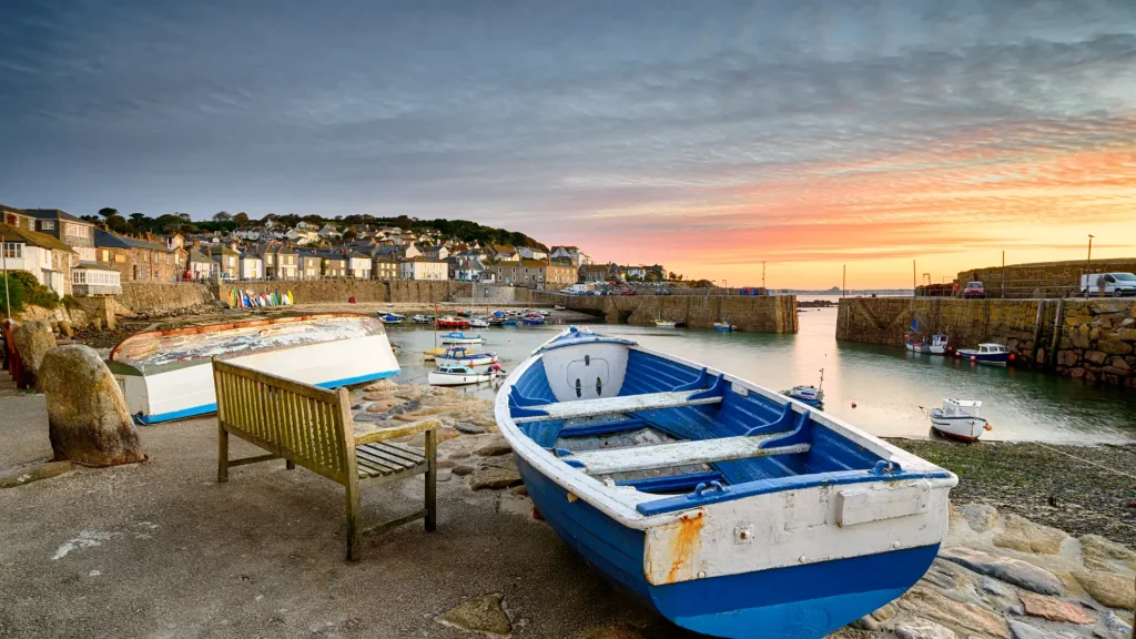 Scenic coastal sunset at a harbor, featuring small boats and a wooden bench in the foreground, with quaint stone buildings in the background.